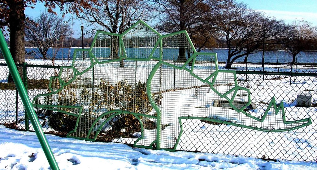 Pale green stegosaurus of bent pipe set into a snowy waterfront park fence